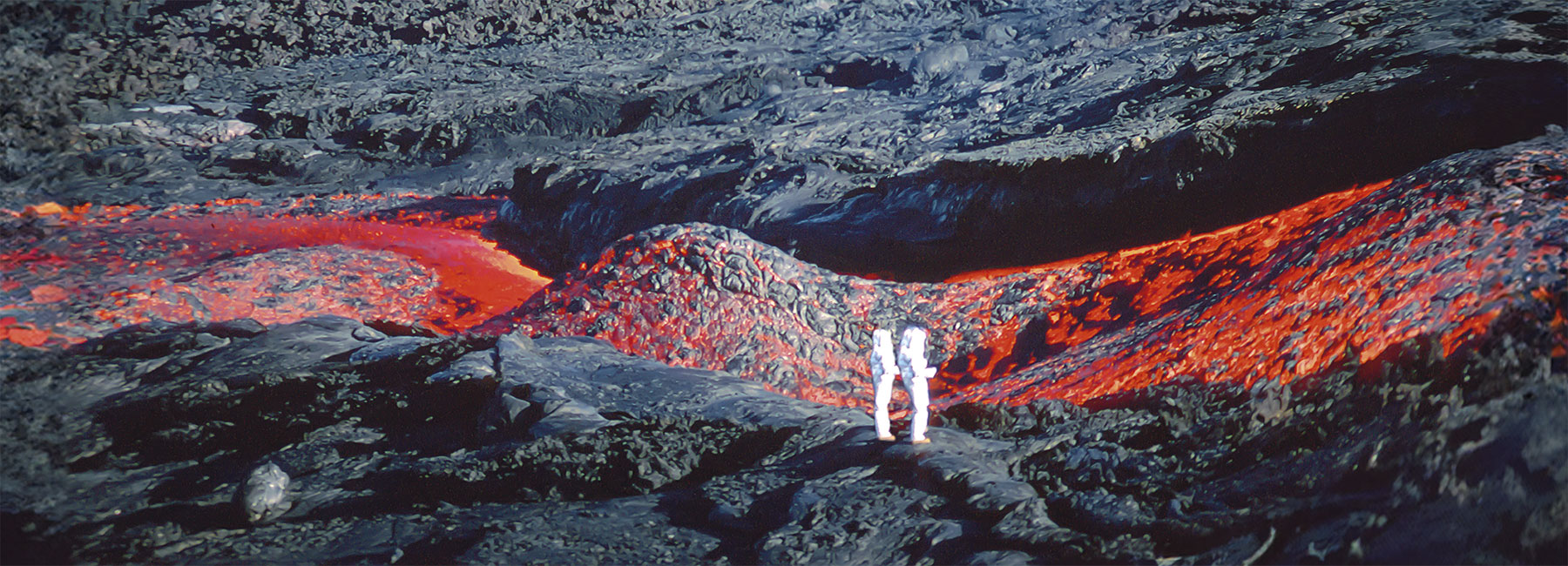 Deux personnes en tenue blanche, se tiennent proche d'une coulée de lave sur une terre magmatique.