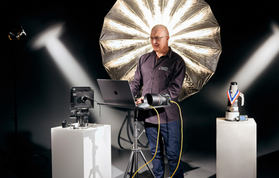 Alain Martineau se tient debout, devant un ordinateur. Il est éclairé par des lumières et entouré d'appareils photographiques.  